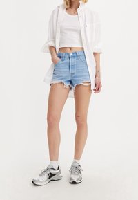 Pantalones cortos de mezclilla azul claro con dobladillo deshilachado, combinados con una blusa corta blanca y una camisa blanca de botones de gran tamaño. Zapatillas deportivas plateadas completan el look.