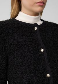 Schwarzer strukturierter Cardigan mit runder Ausschnitt, zwei silbernen dekorativen Knöpfen und einem weißem Rollkragenpullover darunter.