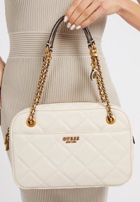 Weiße gesteppte Handtasche mit goldenen Kettenakzenten und einem Logo auf der Vorderseite. Verfügt über zwei Griffe und eine glatte Textur.