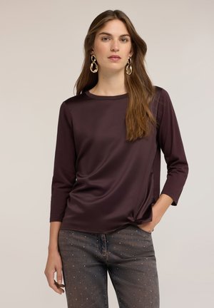 Mujer con cabello largo y castaño que lleva grandes pendientes de aro dorados, una camisa de manga larga morada oscura y vaqueros oscuros con pequeños tachones, con la mano en el bolsillo.
