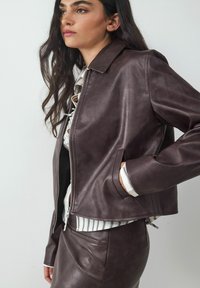 Bruine leren jas met een rits aan de voorkant, puntige kraag en zijzakken; gedragen over een gestreept shirt. Glad textuur en getailleerd ontwerp.