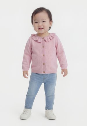 Tout-petit debout, portant un gilet en tricot rose avec un col à volants, un jean bleu clair et des baskets blanches, souriant devant un fond uni.