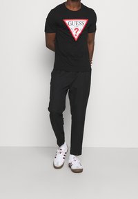 Muž v černém tričku Guess s červeným trojúhelníkovým logem, černých kalhotách a bílých teniskách s červenými pruhy, stojící na bílém pozadí.