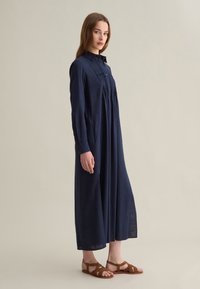 Lungo abito in lino blu navy con colletto, apertura con bottoni e dettagli plissettati; abbinato a sandali marroni con cinturini.