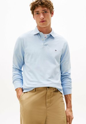 Light blue long-sleeve polo shirt with a collar, featuring a small logo on the chest. Paired with khaki-colored trousers.