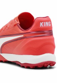 Chaussure de sport rouge avec une tige texturée, des accents blancs et une étiquette à l'arrière indiquant "KING". Caractéristiques d'une semelle en caoutchouc avec des pointes de traction.