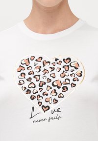 Witte t-shirt met een grote hartafbeelding met roze en zwarte ontwerpen, benadrukt met goud. Onder de afbeelding staat de tekst "Liefde faalt nooit."