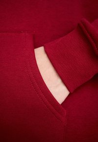Main partiellement dans la poche avant d’un sweat-shirt rouge avec un poignet côtelé au niveau du poignet.