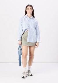 Camicetta blu chiaro a righe con bottoni, gonna beige, calzini bianchi e sneakers grigie. Borsa blu con tracolla stampata. Outfit semplice e casual.