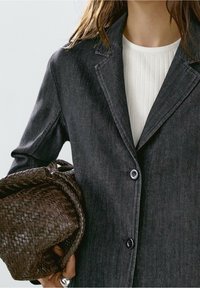 Veste en denim foncé avec col cranté, deux boutons, et un sac brun tissé texturé tenu dans une main. Haut blanc côtelé en dessous.