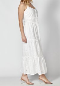 Vit maxiklänning med ögonhålspattern, justerbara tunna remmar, flerlagrig volangslut och knappdetaljer fram. Bärs med klara klacksandaler.