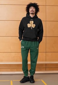 Sweatshirt preta com capuz e design de trevo dourado, com bolso frontal, combinada com calças de fato de treino verdes com texto branco "Dawn City" e sapatilhas pretas.