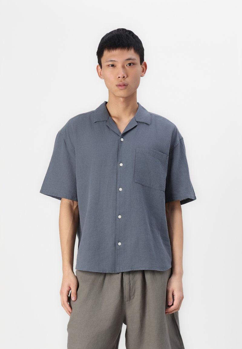 Chemise à manches courtes avec col en tissu gris doux, dotée d'une poche avant et d'une texture subtile. Associée à un pantalon gris clair.