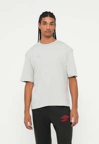 T-shirt cinza claro de manga curta com um corte solto; apresenta um logotipo discreto no peito. Usado com calças pretas com bordados vermelhos.