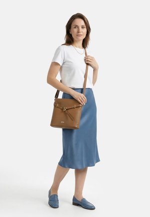 Vrouw draagt een wit T-shirt, een blauwe rok tot op de knie, blauwe loafers, en houdt een bruine leren schoudertas met gouden gesp vast.