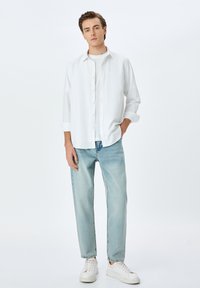 Camisa blanca de botones sobre una camiseta blanca, combinada con jeans azul claro y zapatillas blancas. Diseño simple y casual con mangas largas.