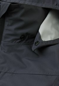 Ciemnoszara tkanina kurtki o gładkiej teksturze, z boczną kieszenią zapinaną na guzik oraz wyściółką z siateczki.