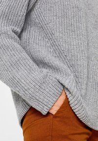 Pull en maille gris clair avec une texture côtelée, doté d'une coupe ample et d'une poche latérale, assorti à un pantalon de couleur rouille.