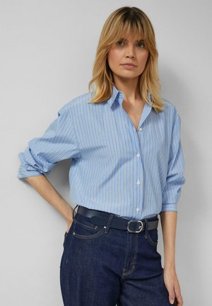 Hellblaues Hemd mit weißen Streifen und kleinen Punkten, lockerer Schnitt, hochgekrempelte Ärmel, kombiniert mit dunklen Jeans und einem blauen Gürtel.