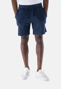 Pantaloni sportivi blu navy con vita elastica e cordoncino, dotati di tasche e realizzati in un morbido tessuto misto cotone. Indossati con scarpe da ginnastica bianche.