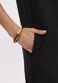 Złotego koloru kanciasta bransoleta noszona na ręce, oparta na czarnej tkaninie kieszeni odzieży, ukazująca elegancki materiał i gładką teksturę.