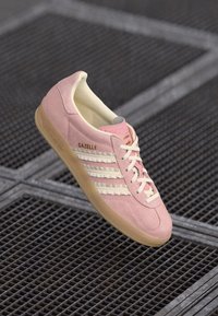 Roze suède sneaker met drie witte strepen, crèmekleurige binnenvoering en gouden "Gazelle" branding. Rubberen gumzool. Klassiek low-top ontwerp.