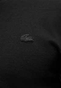 Μικρό μαύρο λογότυπο κροκόδειλου Lacoste, κεντημένο σε μαύρο ύφασμα ενδύματος, που δείχνει λεπτομερή υφή και ραφή.
