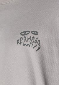 Light gray textured fabric t-shirt featuring a black graphic print with abstract eyes and the word "KARMA8".