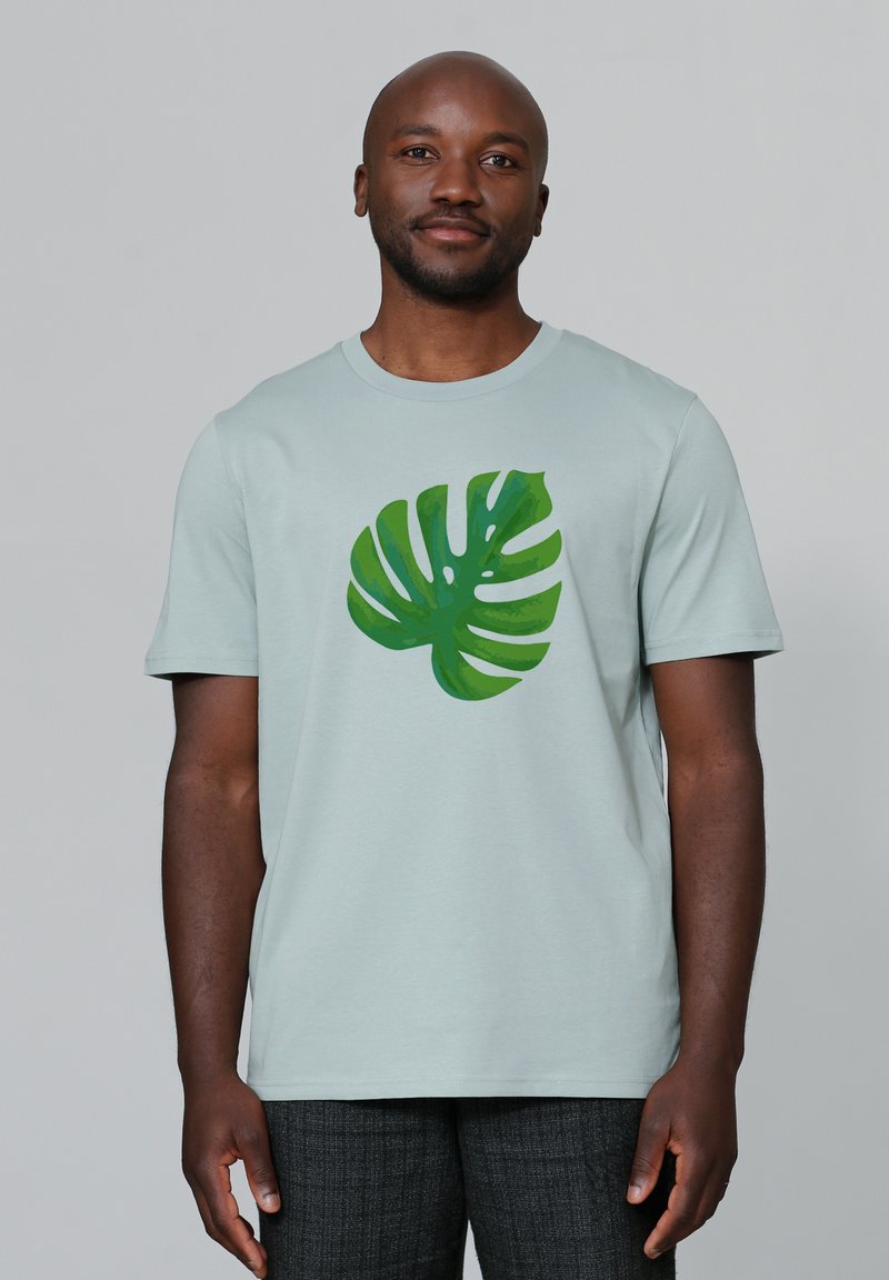 T-shirt en coton vert clair avec un grand graphique de feuille de monstera verte sur le devant. Manches courtes et design col rond.