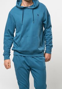 Modrá mikina s kapucí vyrobená z měkké látky, má klokaní kapsu, šňůrkovou kapuci, žebrované manžety a malé logo na hrudi.