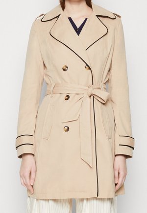 Trench-coat beige à double boutonnage avec liseré noir, boutons effet écaille, ceinture à nouer à la taille et pattes aux poignets, porté sur un pantalon à rayures.