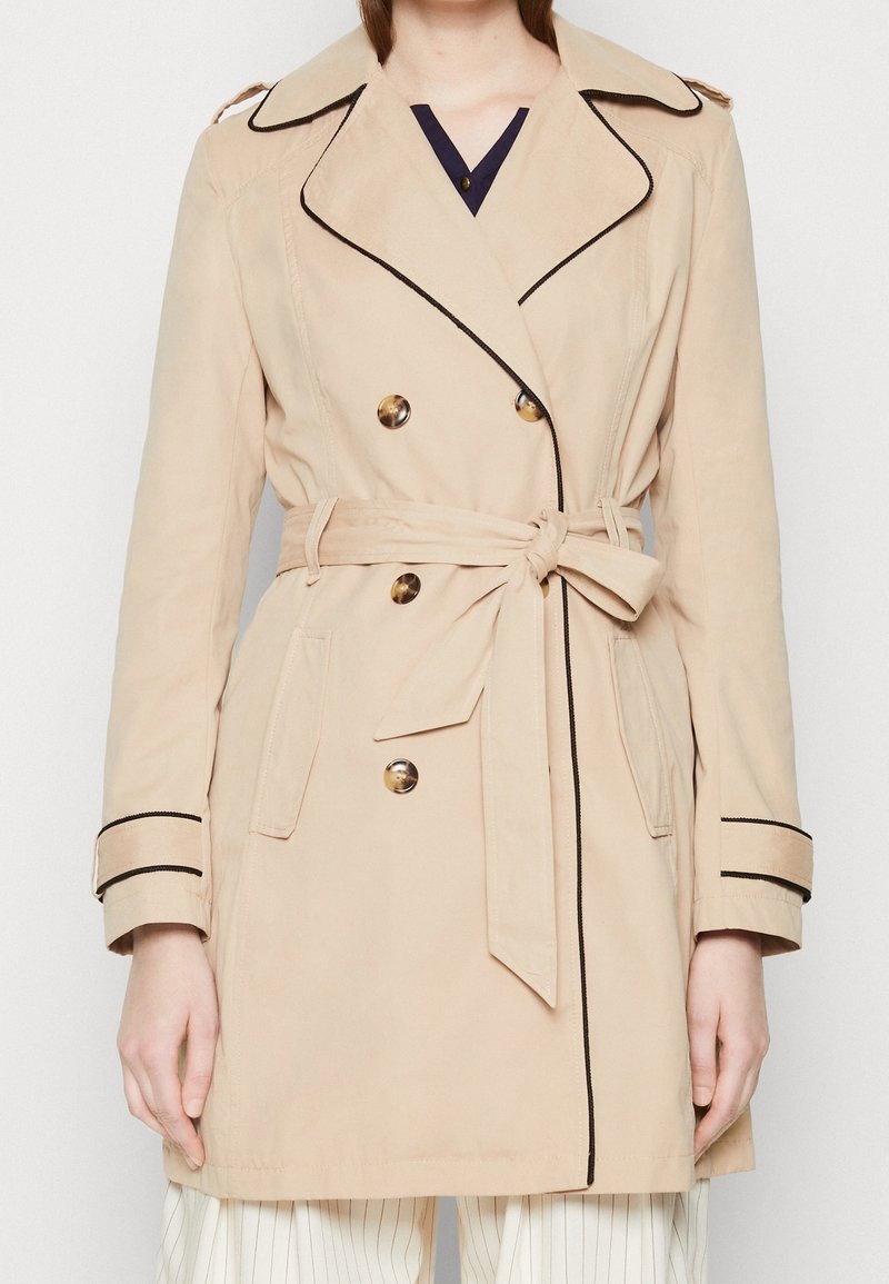 Trench-coat beige à double boutonnage avec liseré noir, boutons effet écaille, ceinture à nouer à la taille et pattes aux poignets, porté sur un pantalon à rayures.