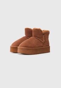 Et par brune ruskinn ankle boots med deilig fôr, glidelås på siden og tykk flat såle på hvit bakgrunn.