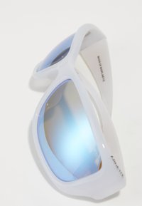 Lunettes de soleil en plastique blanc avec des verres miroités bleus, dotées d'un design enveloppant et d'une texture lisse. Marquage visible sur le côté.