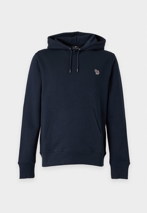 Mężczyzna w granatowej bluzie z kapturem z małym emblem, szare plisowane spodnie i czarne buty, stoi z rękami w kieszeniach na jasnym tle.