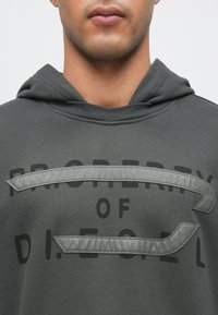 Sivi pulover s kapuco z rebristim izrezom in velikimi črnimi črkami. Posebnost je mehak sivi tkaninski detajl čez prsi.