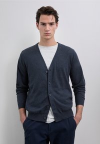 Cardigan gris foncé en maille, avec un col en V, une fermeture à boutons et des poignets côtelés, porté sur une chemise gris clair.