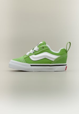 Baskets en toile verte avec lacets blancs, bande latérale ondulée blanche, semelle en caoutchouc blanche et tirette au talon sur un fond neutre.