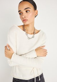 Jeune femme aux cheveux coiffés en arrière, portant un pull en maille crème, un collier chaîne argenté oversize et de petites boucles d'oreilles créoles, les bras croisés.