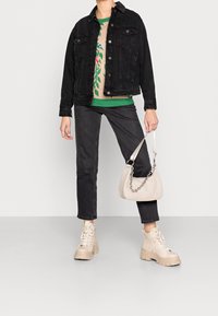 Veste en denim noir, pull beige avec des accents verts, jeans foncés et bottes épaisses beiges. Tenant un petit sac beige avec une bandoulière en chaîne.