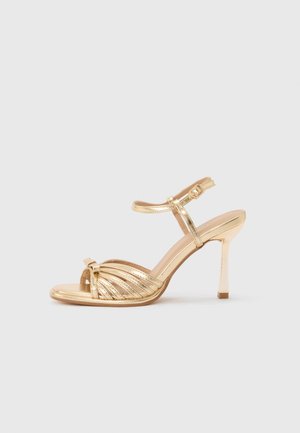 Sandale à talon haut en or avec bout ouvert, plusieurs brides à l'avant, et bride à boucle autour de la cheville sur un fond neutre.