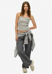 Grijze cropped tanktop met tekstdetail, gecombineerd met losse grijze broek en een gestreepte shirt geknoopt om de taille. Sneakers in neutrale tinten.