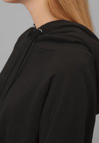 Czarna bluza z kapturem o gładkiej teksturze, wyposażona w sznurek i okrągły dekolt, zaprezentowana z boku na neutralnym tle.