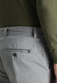 Pantaloni grigi con una texture liscia, dotati di una tasca posteriore con chiusura a bottone e un accento con logo, abbinati a una maglietta verde oliva.