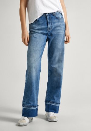 Person mit blauen Jeans mit weitem Bein und umgeschlagenem Saum, weißen Sneakers und einem in die Hose gesteckten weißen T-Shirt vor einem schlichten hellen Hintergrund.