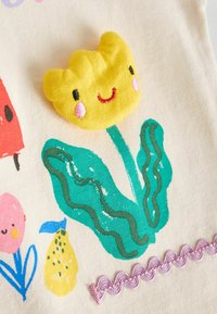 Textil brodat în culori multiple, prezentând o floare galbenă zâmbitoare, modele de frunze verzi, margini roz în formă de scoică și diverse desene florale vibrante.