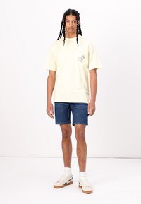 T-shirt en coton jaune avec une poche sur le devant et un design graphique, associée à un short en denim bleu et des baskets blanches.