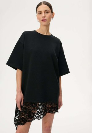 Kobieta o ciemnych włosach związanych w niski kok, ubrana w oversize'ową czarną sukienkę T-shirt z asymetrycznym koronkowym dołem, stojąca na jednolitym jasnym tle.