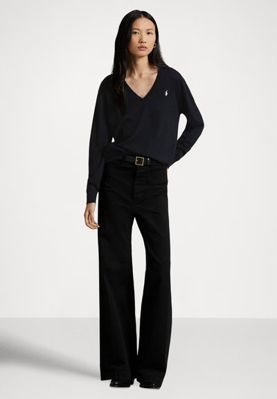 Black v-neck sweater, long sleeves, featuring a small logo on the chest. Paired with high-waisted black wide-leg pants and a black belt.