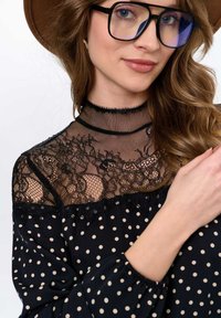 Zwarte blouse met stippenpatroon en kantoverlay, hoge hals en ruches. Model draagt grote bril en een bruine hoed. Zachte stof.